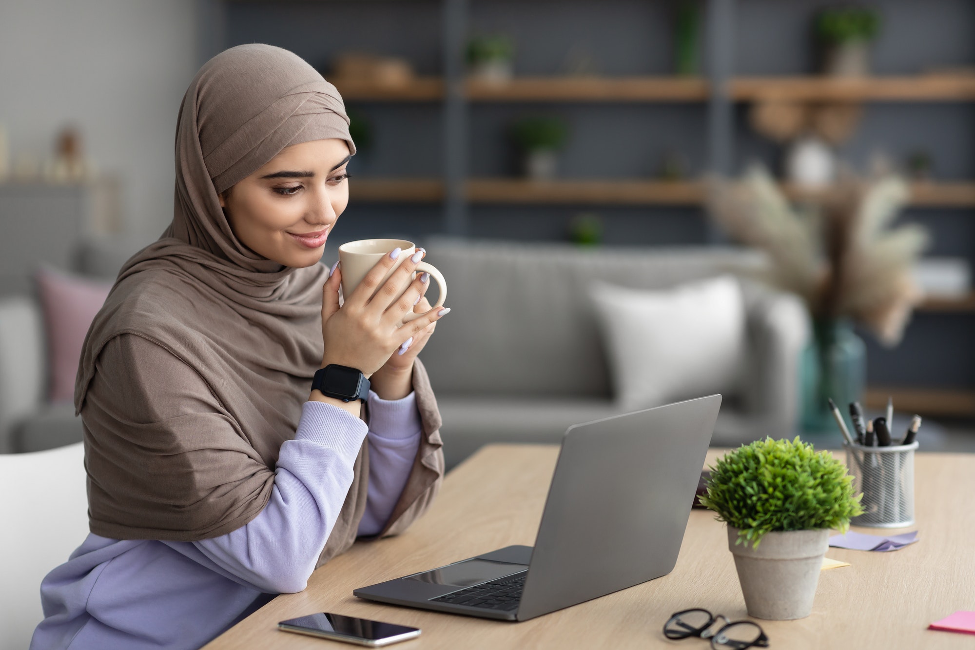 Muslim lady watching video on pc, holding cup drinking coffee