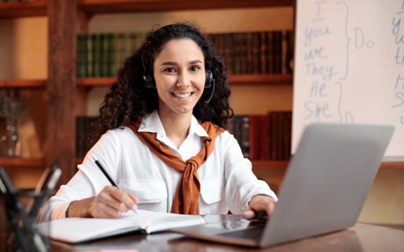 Lady sitting at table, using computer and writing in notebook