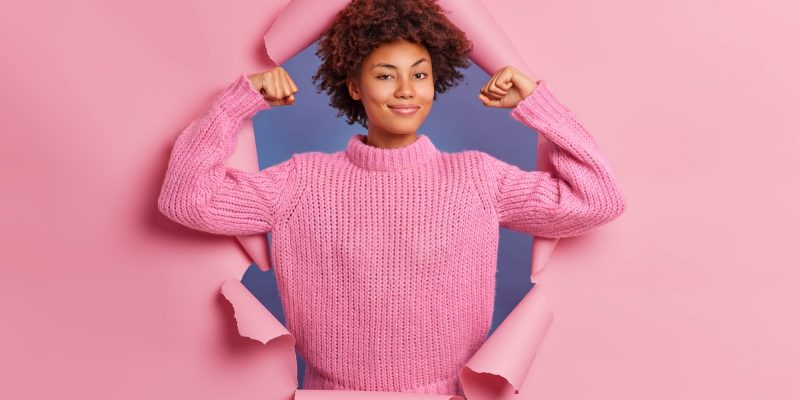 Self assured beautiful young Afro American woman raises arms shows biceps being strong and powerful